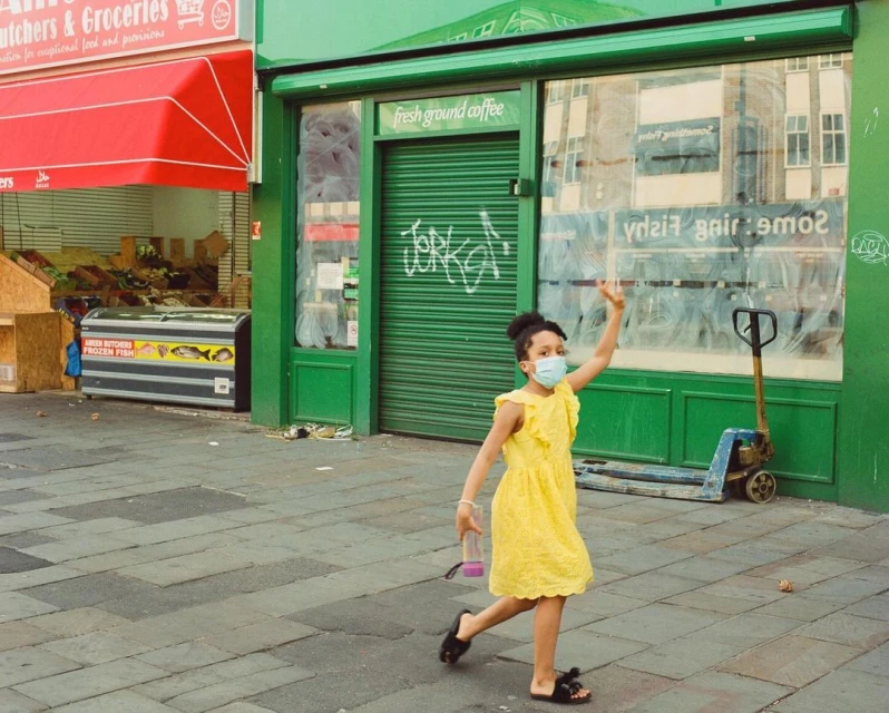 Young girl in a yellow dress and mask waves while walking on a city sidewalk with shops and buildings in the background on a sunny day.