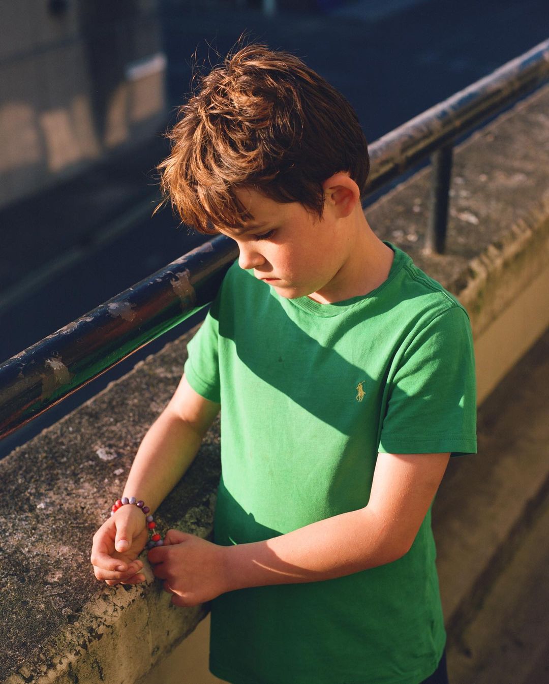 A young boy in a green t-shirt stands outside with his head down, wearing beaded bracelets, on a sunny day with shadows.