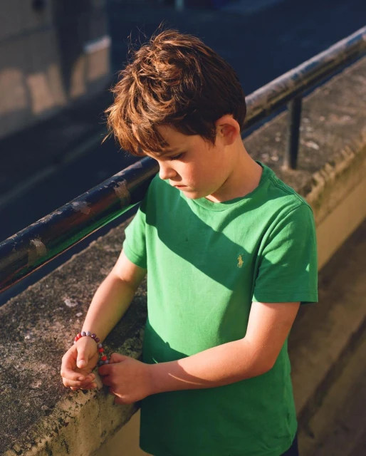 A young boy in a green t-shirt stands outside with his head down, wearing beaded bracelets, on a sunny day with shadows.