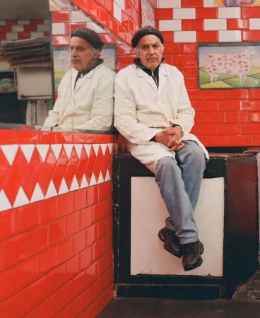 An older man in a white butcher coat and dark cap sits against a red and white tiled wall, reflected in a large mirror.