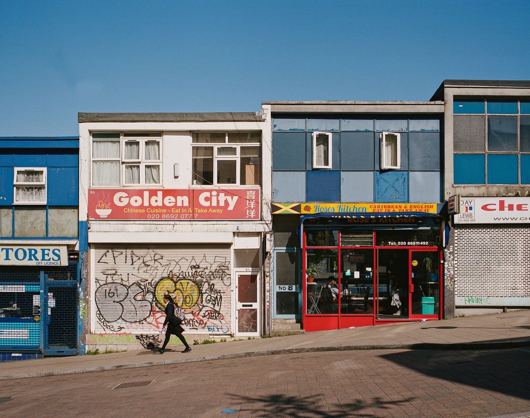 A person walks past shops in an urban area during daytime, with colorful storefronts and clear blue sky above the buildings.