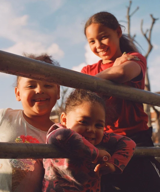 Three smiling children pose behind metal bars in a sunlit outdoor setting with a blue sky. Playful childhood, joyful expressions, family. 