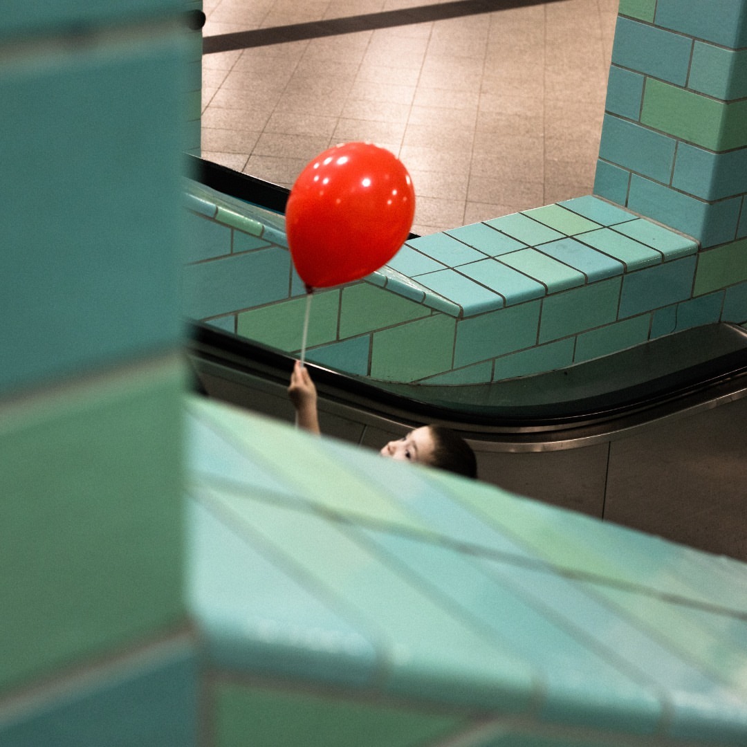 Child holds a red balloon while riding up an escalator next to tiled walls, creating a sense of whimsy and movement in an urban setting.