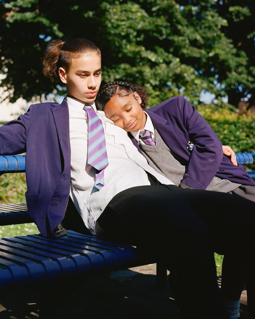 Two teenagers in school uniforms sit on a park bench on a sunny day. One rests their head on the other's shoulder. Lush trees in background.