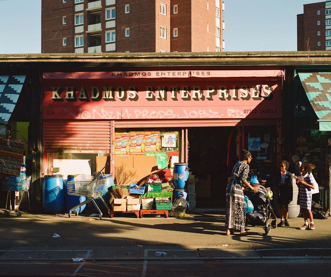 Family stands outside Khadmus Enterprises store with a red awning and exterior. Barrels, boxes, and a shopping cart sit nearby.