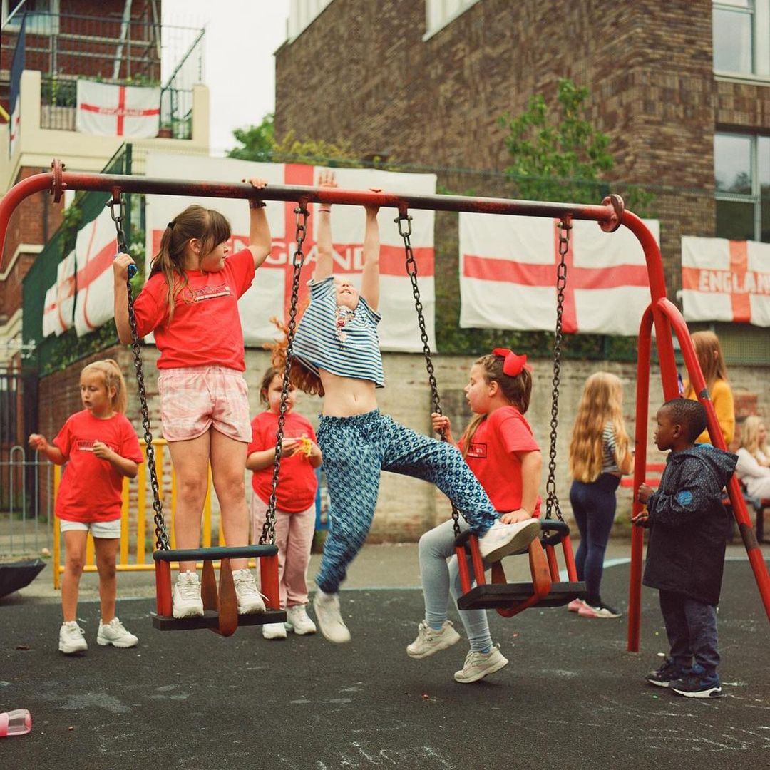 Children enjoy a playground with red metal swings and climbing equipment. Several kids wear matching red shirts. English flags hang above.