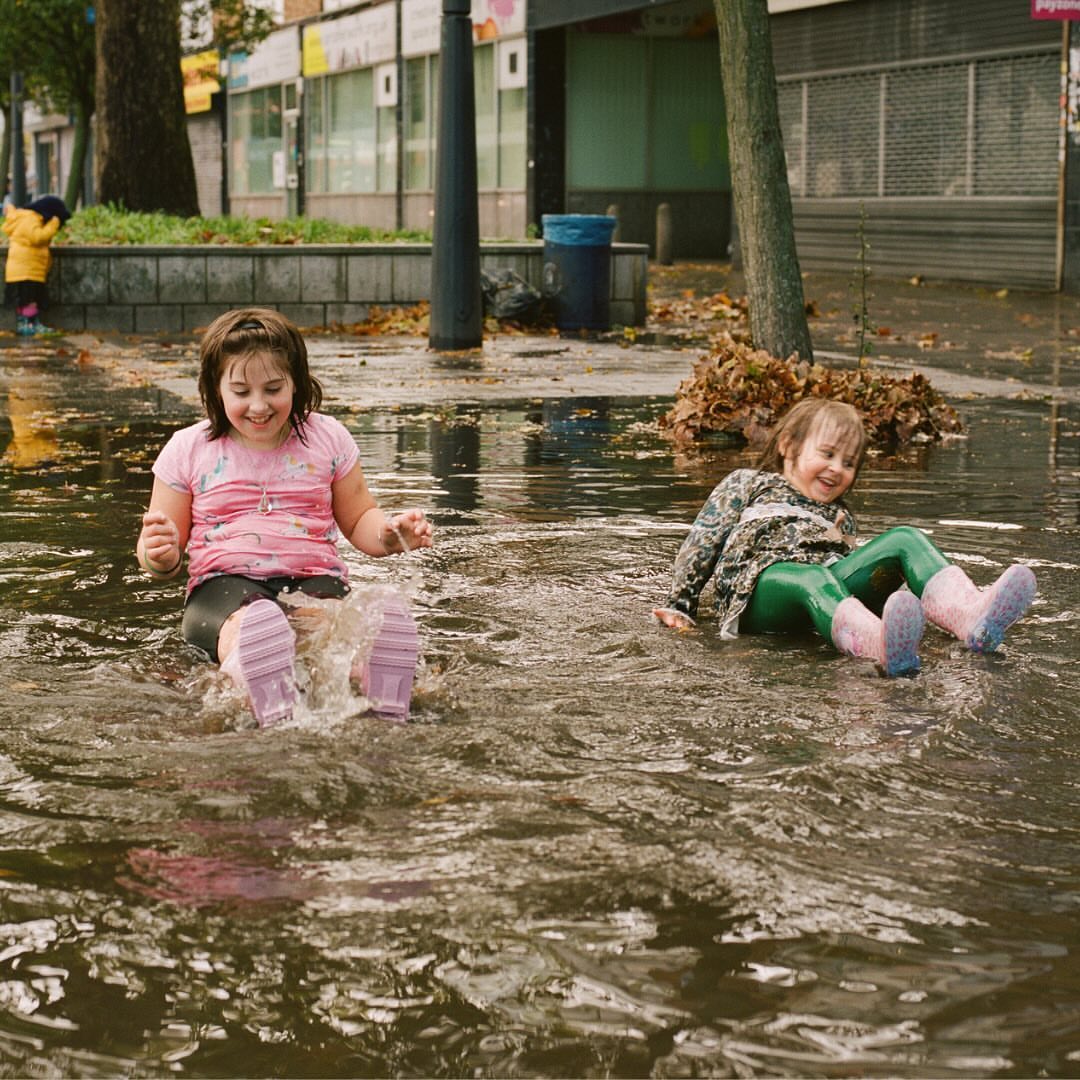 Two young girls in pink boots play in a flooded street, gleefully splashing water; one girl wears a pink top, the other a patterned jacket.