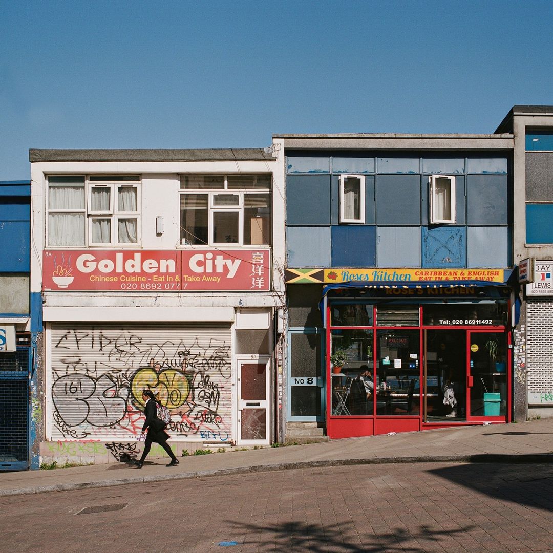 A woman walks past a row of colorful buildings in an urban setting. The buildings feature storefronts with signage and graffiti.