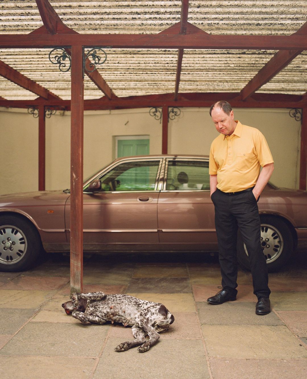 Adult male in a yellow shirt standing next to a tan car with a spotted dog lying on the ground near a building with a covered patio.