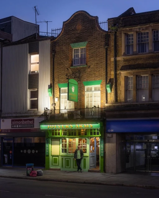 A man stands outside the Joiners Arms pub at night. The brick building is lit by interior and exterior lights, casting warm tones.