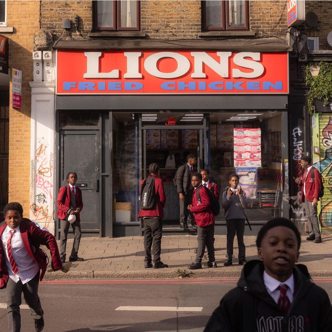 Several African-descent children in uniform standing in front of Lions Fried Chicken in urban setting, some kids are walking on the street.