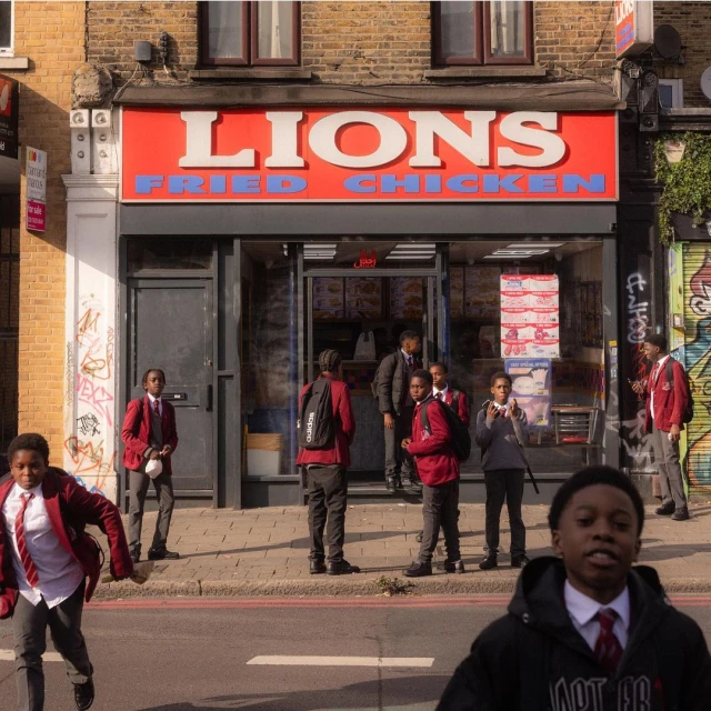 Several African-descent children in uniform standing in front of Lions Fried Chicken in urban setting, some kids are walking on the street.