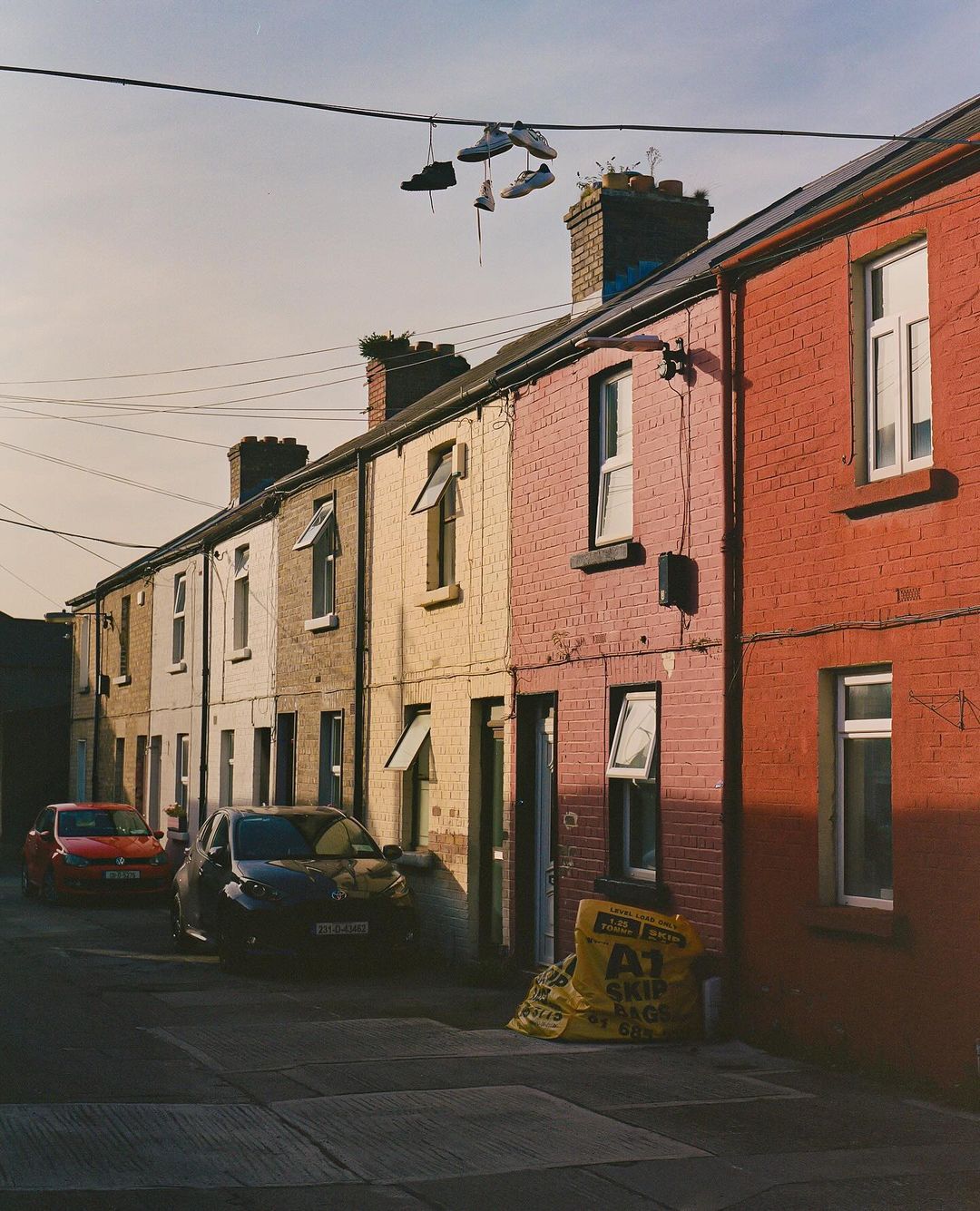 Street with colorful houses and shoes hanging from a wire in front, two cars, and yellow skip bags.