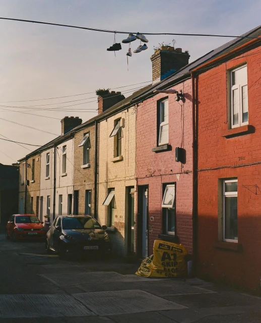 Street with colorful houses and shoes hanging from a wire in front, two cars, and yellow skip bags.