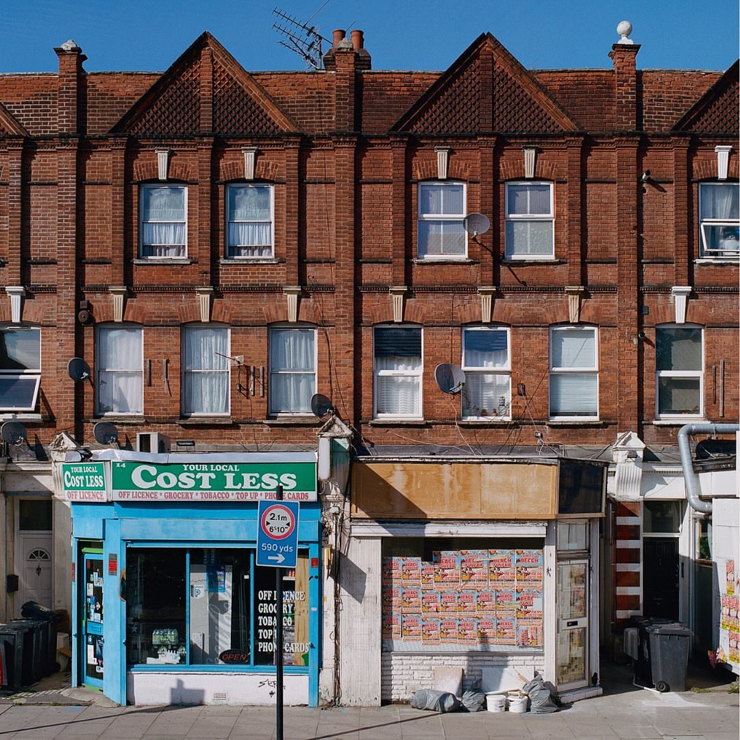 Two-story brick building with small shops on the ground floor, featuring blue, green, and white facades, set against a clear blue sky.