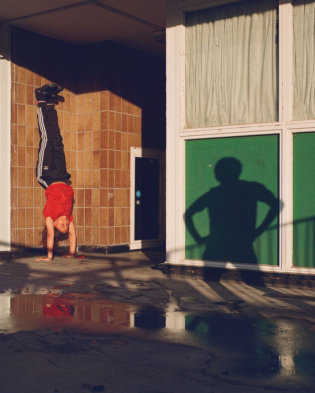 Young boy performs a handstand against a tile wall in the sunlight; his shadow is visible on a nearby window with curtains.