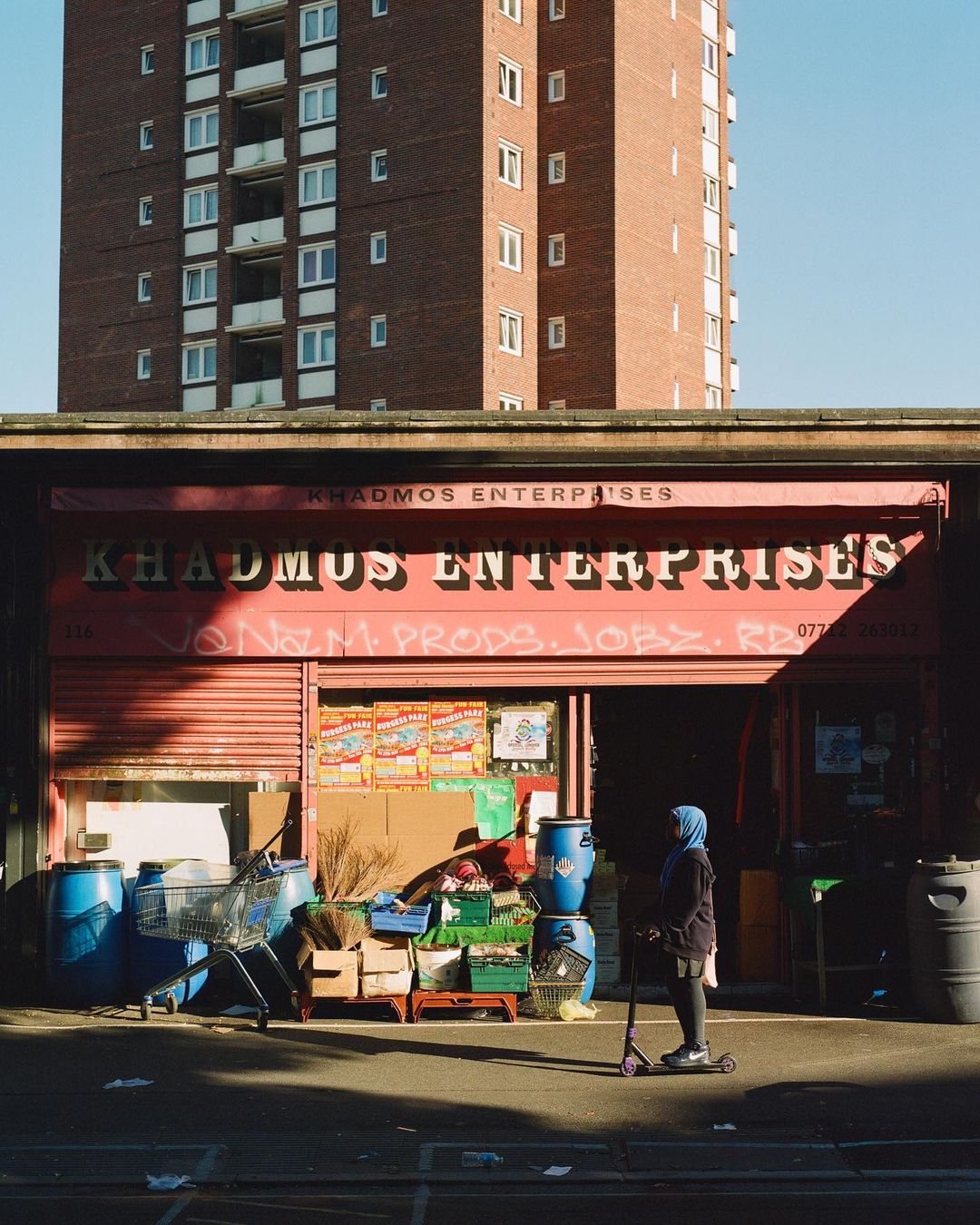 Woman in hijab on scooter outside Khadmus Enterprises with blue barrels, shopping cart, and apartment building backdrop in urban scene.