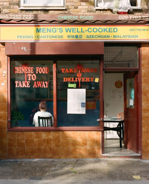 The facade of Meng's Well-Cooked Chinese food restaurant, featuring large windows, takeout sign, with a woman seated inside.
