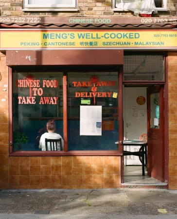 The facade of Meng's Well-Cooked Chinese food restaurant, featuring large windows, takeout sign, with a woman seated inside.