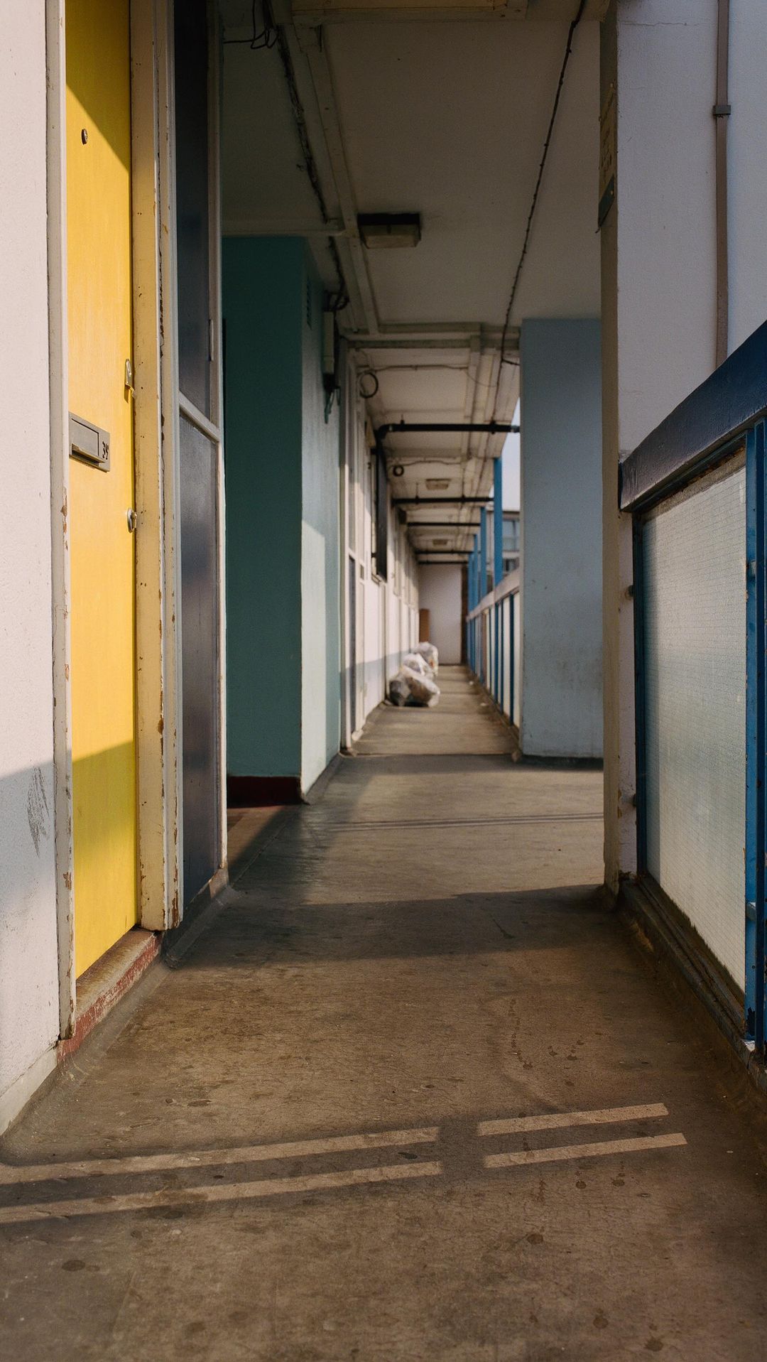Residential building walkway featuring yellow doors, concrete floors, and an open-air design with visible pipework and natural light.