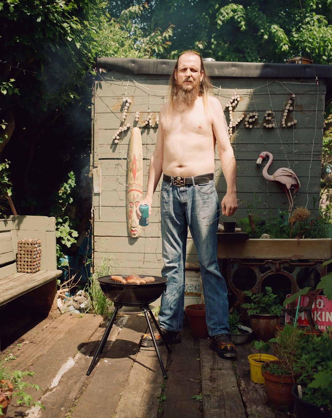 An adult male stands shirtless next to a barbecue in a garden setting, with a shed decorated with shells and plants in the background.