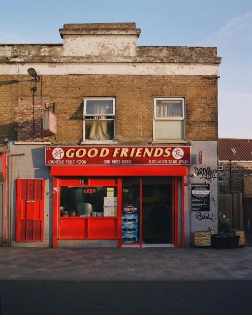 Red storefront of the Good Friends Chinese fast food restaurant in a worn brick building on a city street with a sunny sky.