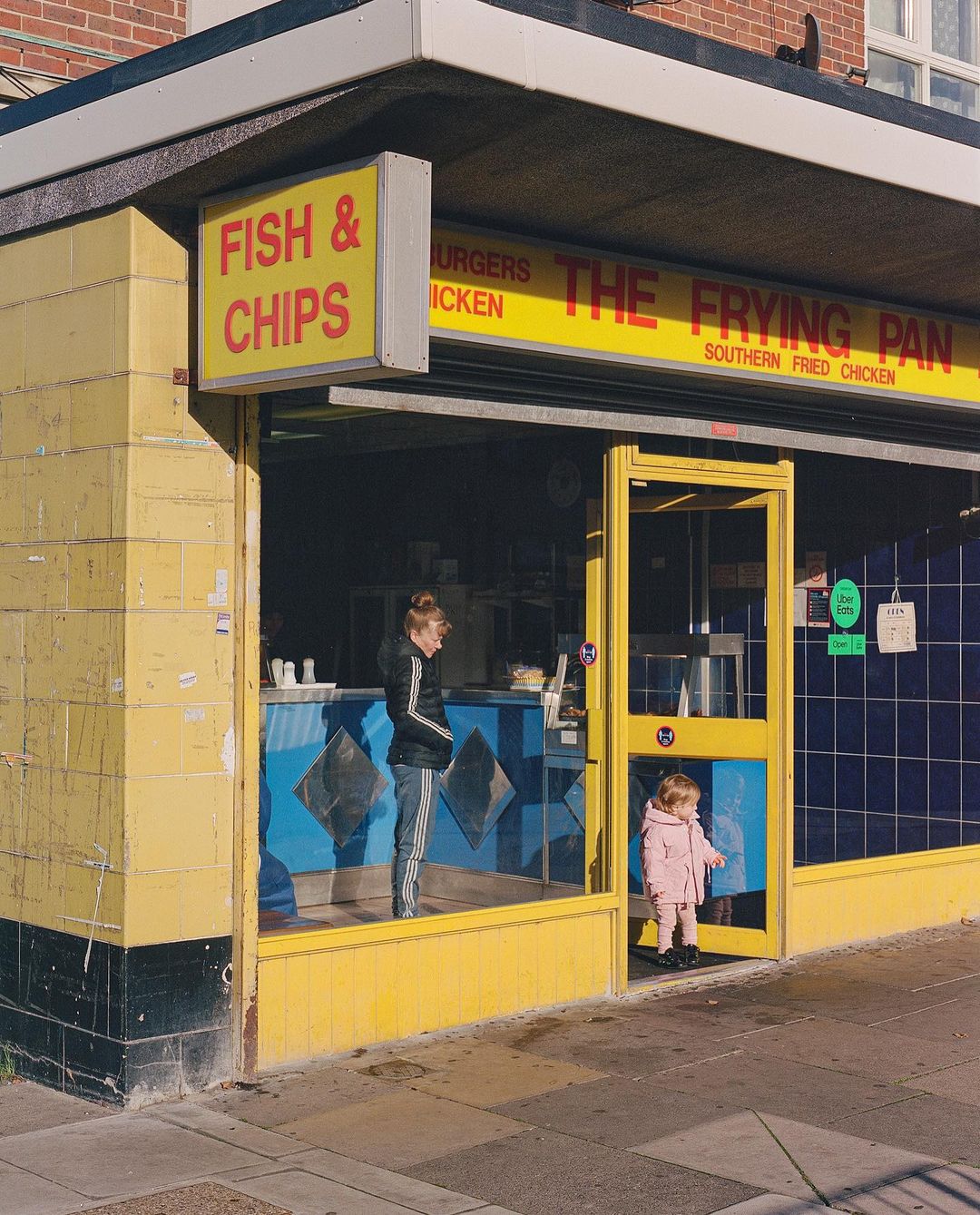 Exterior view of The Frying Pan fish and chips shop on a sunny day, featuring a woman and child at the entrance, London.