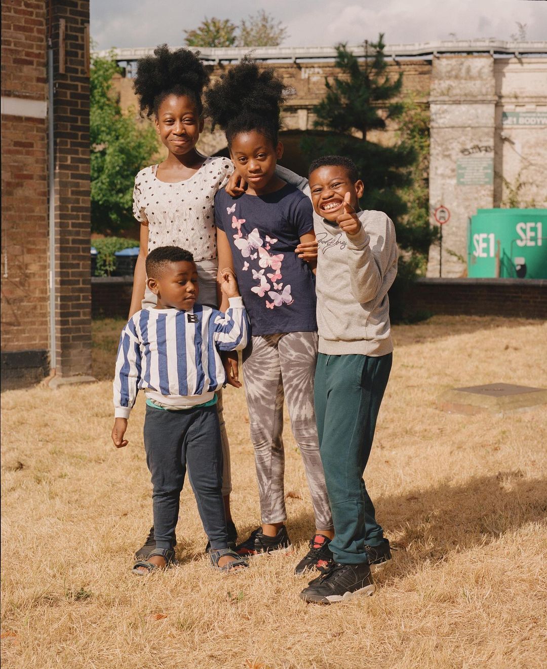 Four smiling children stand on a grassy field near brick buildings and an overpass, creating a cheerful and casual outdoor portrait.