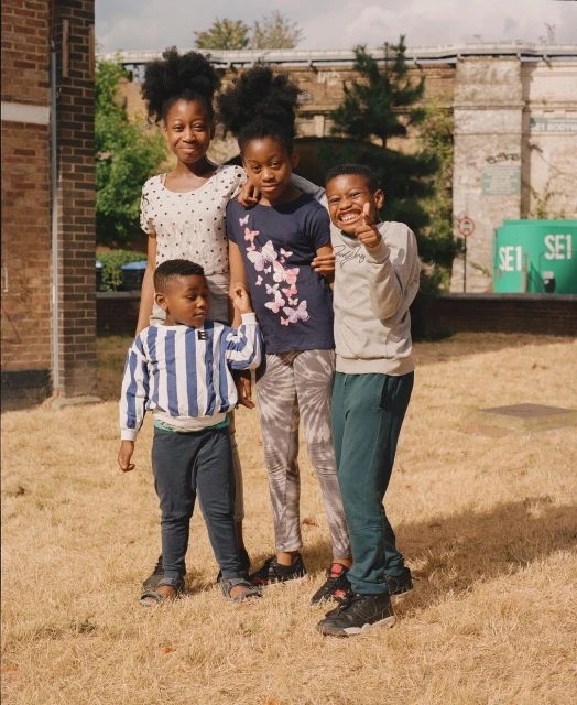 Four smiling children stand on a grassy field near brick buildings and an overpass, creating a cheerful and casual outdoor portrait.
