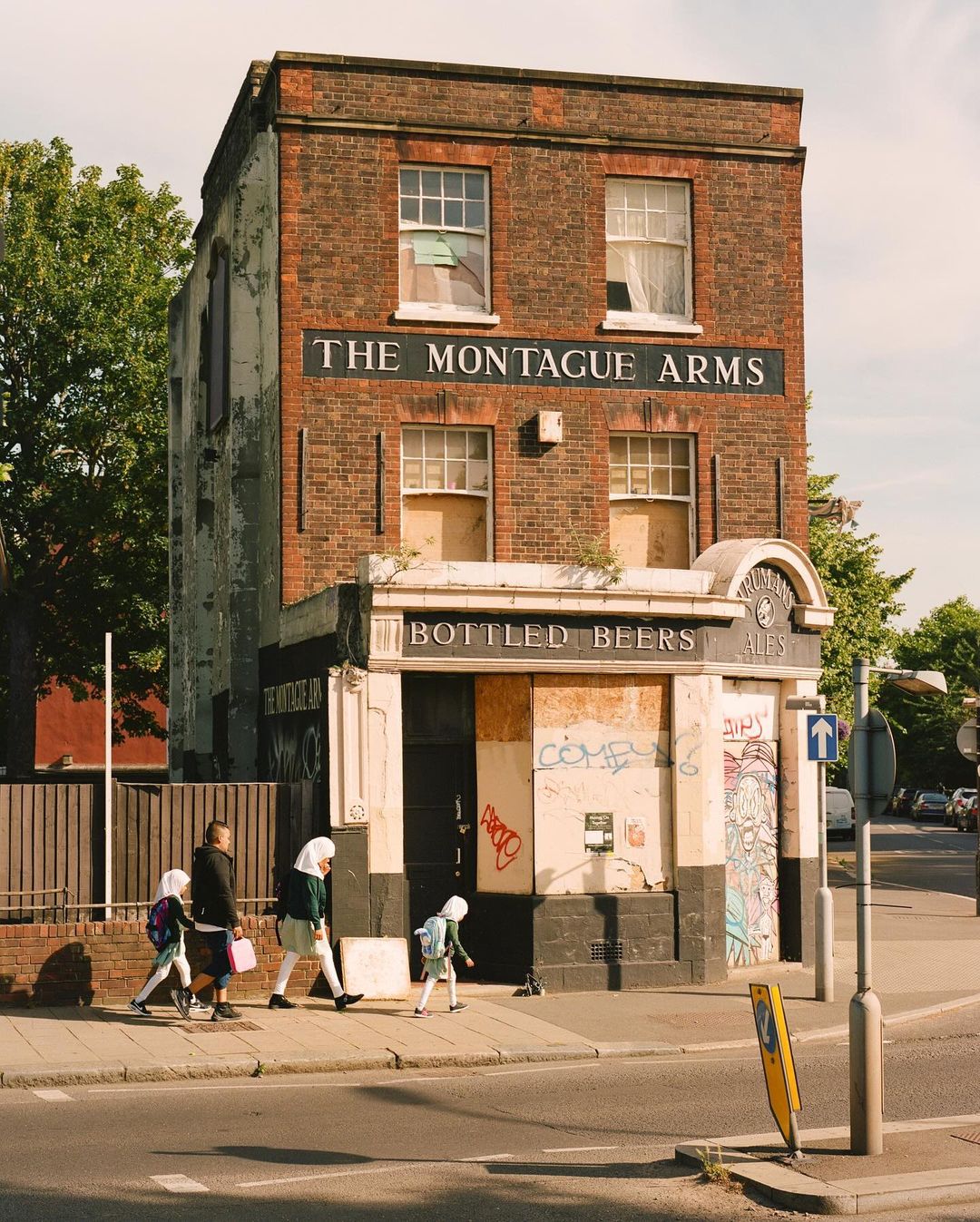 Urban scene shows group crossing street in front of boarded-up brick building with 'The Montague Arms' sign, juxtaposing decay and daily life.