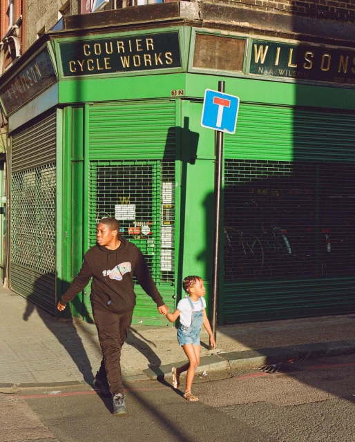 A young adult and a child walk hand-in-hand in an urban environment. The building behind them has a green facade and bold signage.