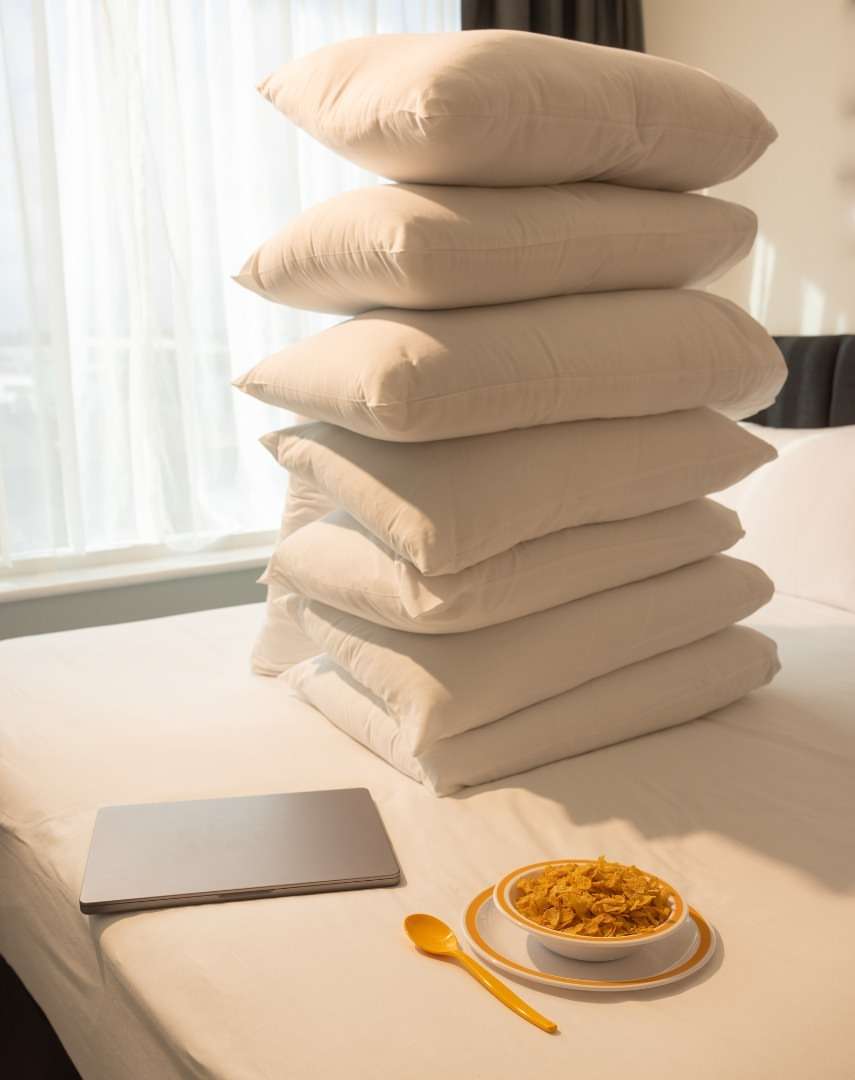 Stack of six white pillows on a bed with breakfast cereal in a bowl, a laptop, and a wood spoon on a white bed cover indoors.