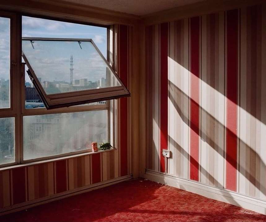 Empty room with red striped wallpaper, open window looking out to city buildings, red carpet, and a can on the windowsill.
