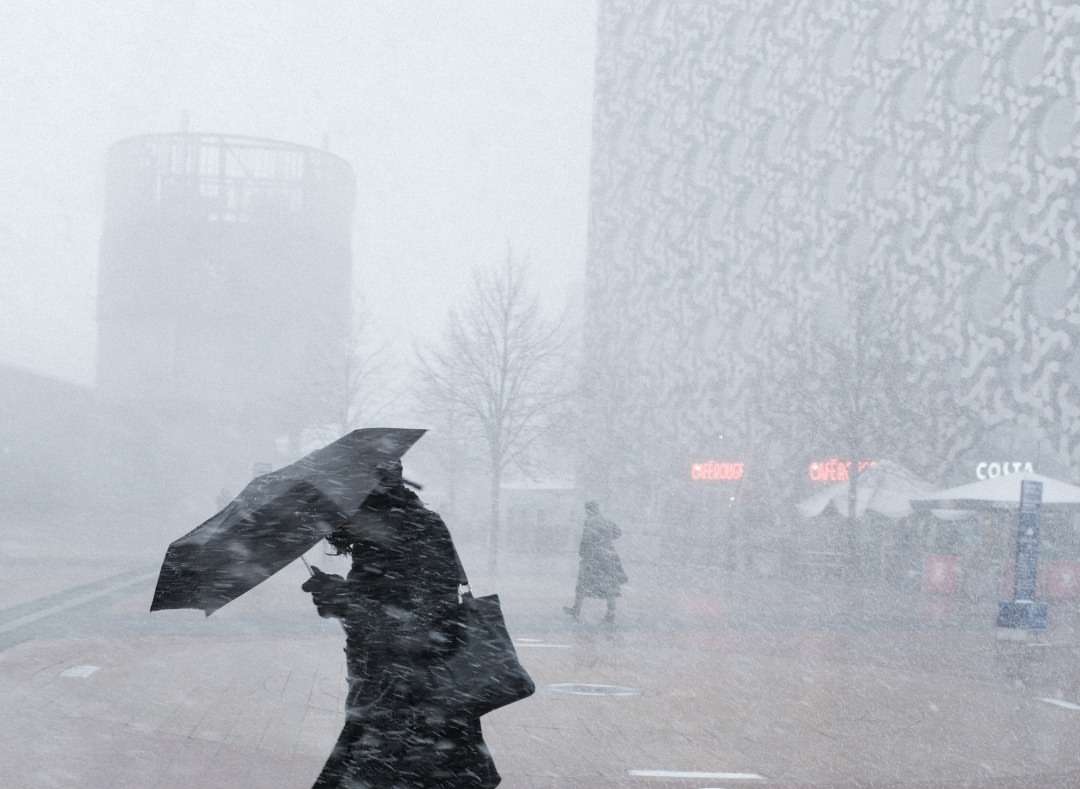 Adult female with umbrella walks through a snowstorm in front of modern buildings, creating a wintry urban scene with muted colors.