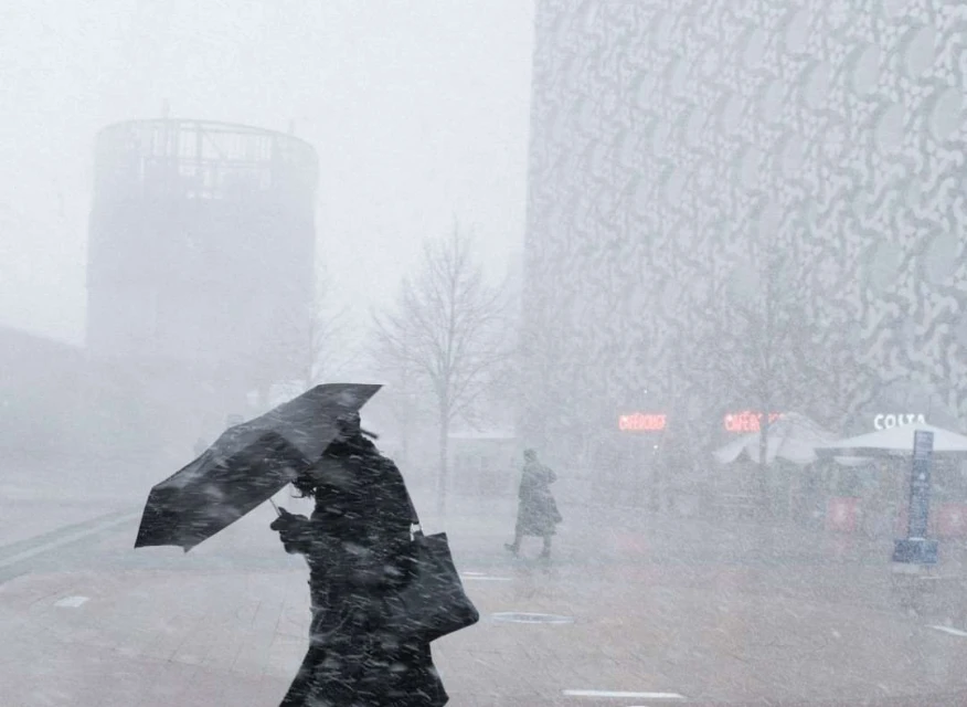 Adult female with umbrella walks through a snowstorm in front of modern buildings, creating a wintry urban scene with muted colors.