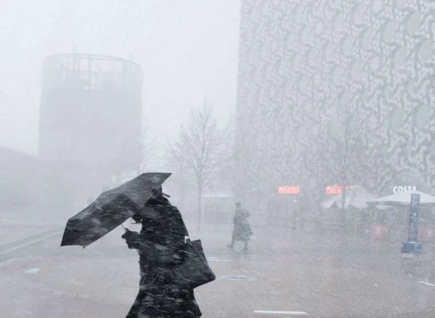 Adult female with umbrella walks through a snowstorm in front of modern buildings, creating a wintry urban scene with muted colors.