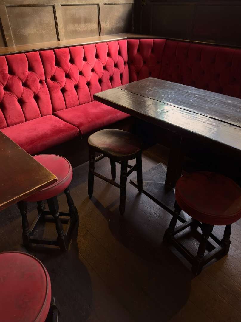 A cozy pub interior featuring dark wooden tables and stools, complemented by a plush, button-tufted red bench and paneled walls.