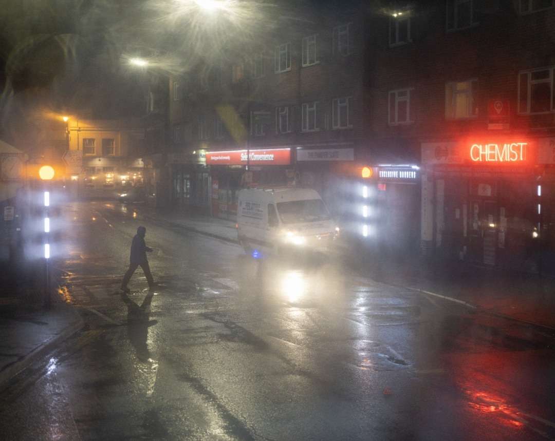 Night scene with a person crossing a wet city street in the rain, illuminated by streetlights and neon signs, creating a hazy effect.