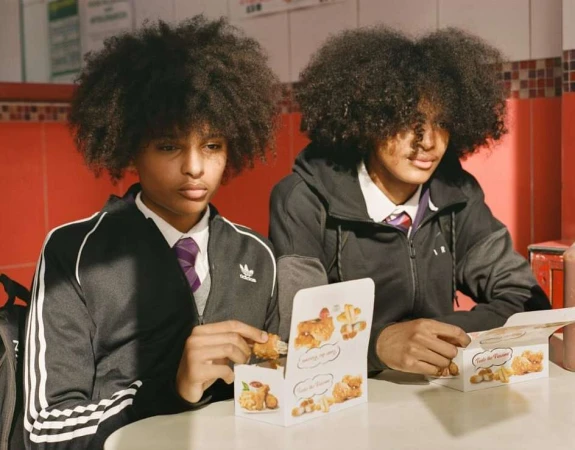 Two Black teenagers in school uniforms sit indoors at a table with fried chicken boxes, enjoying a meal in a casual setting.
