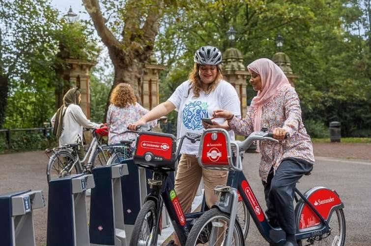 Two women on Santander bikes in a park, one wearing a helmet and the other a hijab, with trees and architecture in background.