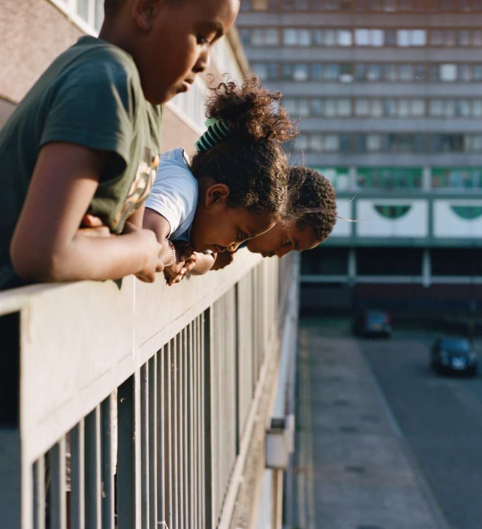 Three children lean on a balcony railing, looking down at a street with cars and a large building in the background, daytime.