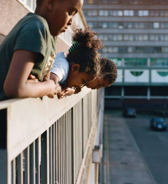 Three children lean on a balcony railing, looking down at a street with cars and a large building in the background, daytime.