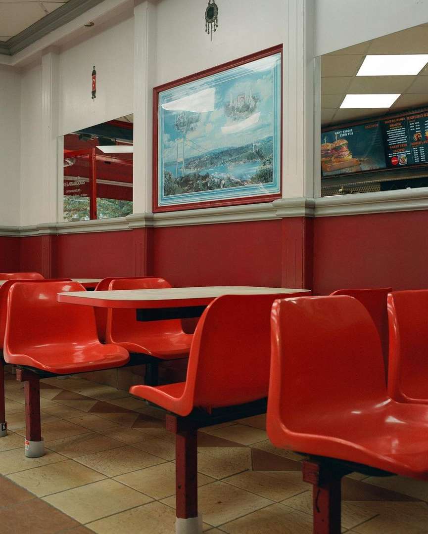Interior of an empty fast-food restaurant with red plastic chairs, off-white tables, and a large landscape painting on the wall.