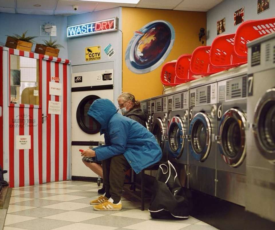 Two people sit inside a laundromat next to rows of washing machines. A person in a blue hooded jacket looks at their phone.