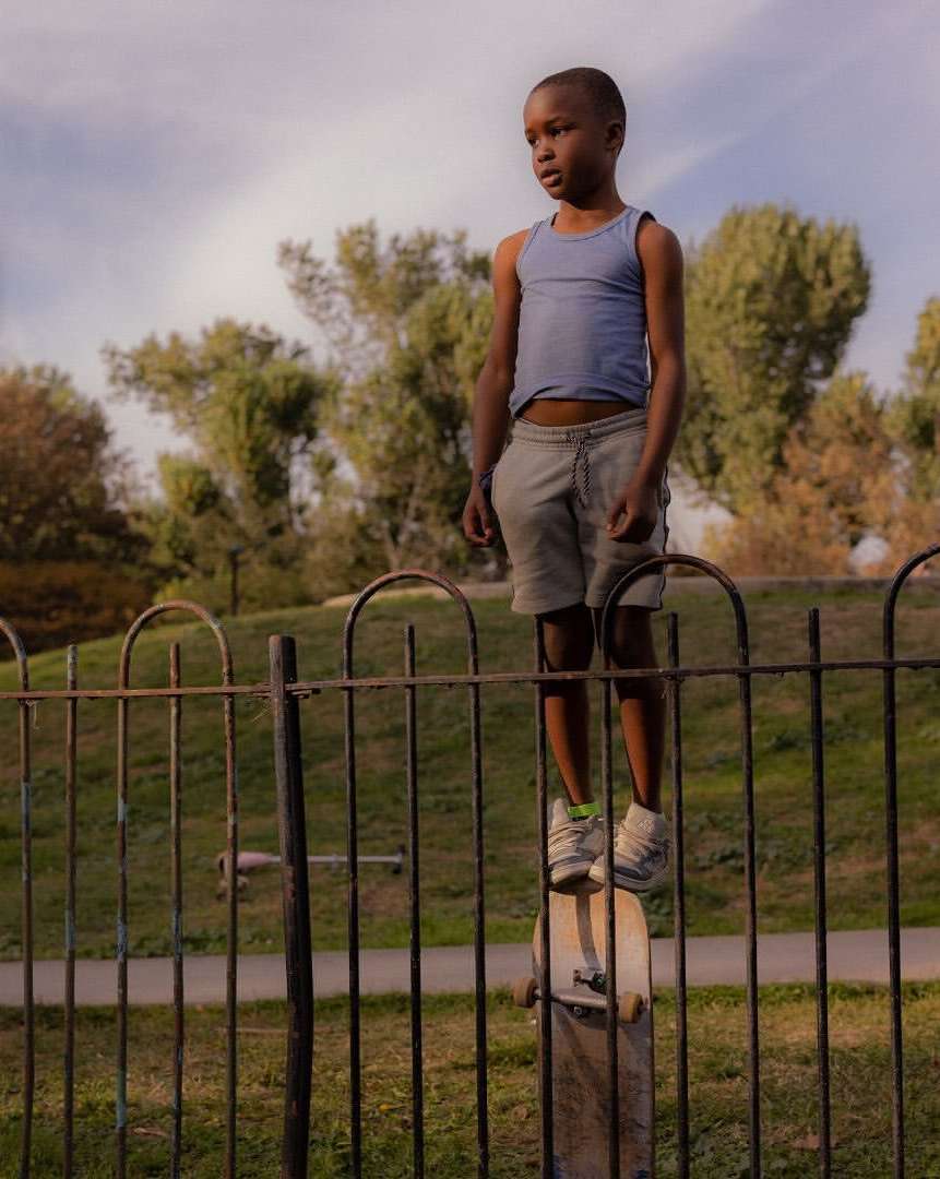 A young black child in a blue tank top stands on a skateboard atop a fence post, framed against a sunny park backdrop with trees.