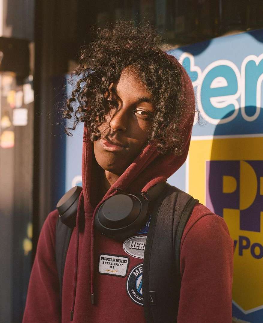 Portrait of a young adult male with curly hair in a maroon hoodie, wearing headphones and a backpack, standing outdoors.