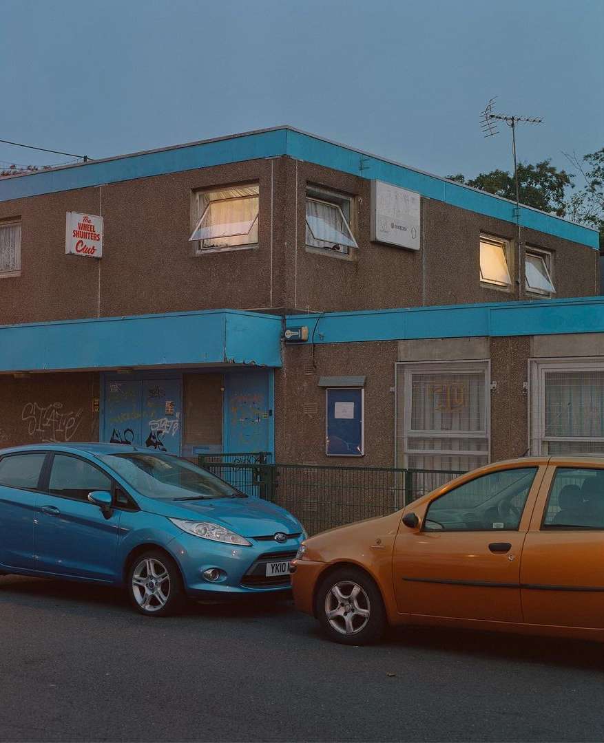 Two cars parked in front of an older apartment building with blue trim, captured during an overcast day with muted lighting.