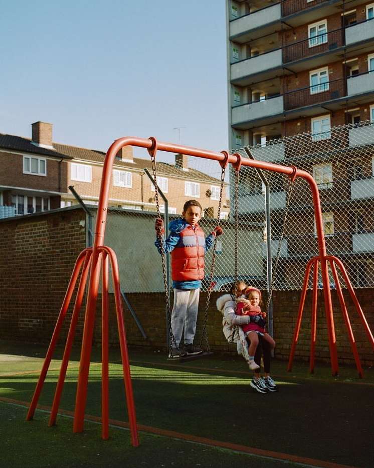 Three children, two girls and a boy, play on a red metal swing set at an outdoor playground with apartment buildings in the background.