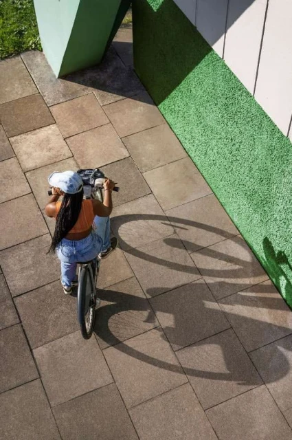 Woman riding a bicycle on a paved surface, captured from an overhead angle with a glimpse of green architecture, casting shadows.