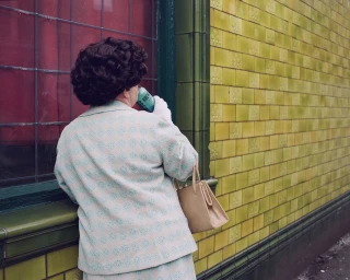 An elderly woman wearing a patterned tweed suit and white gloves drinks from a can outside a pub with yellow tiles.
